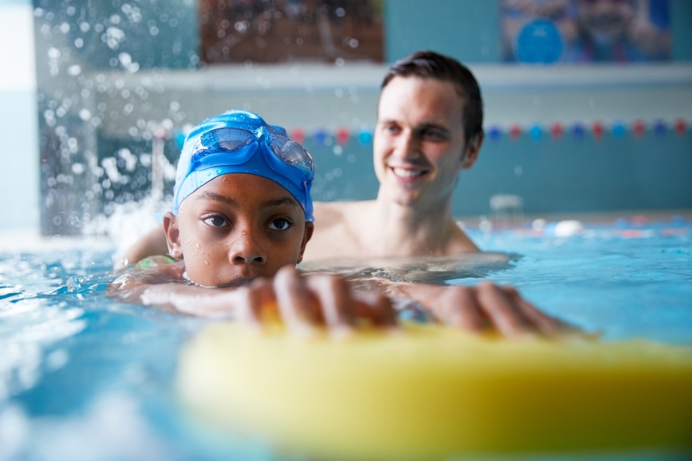 A focused boy in a swim cap and goggles holds onto a floating device in a pool with an instructor standing beside him during swim lessons.