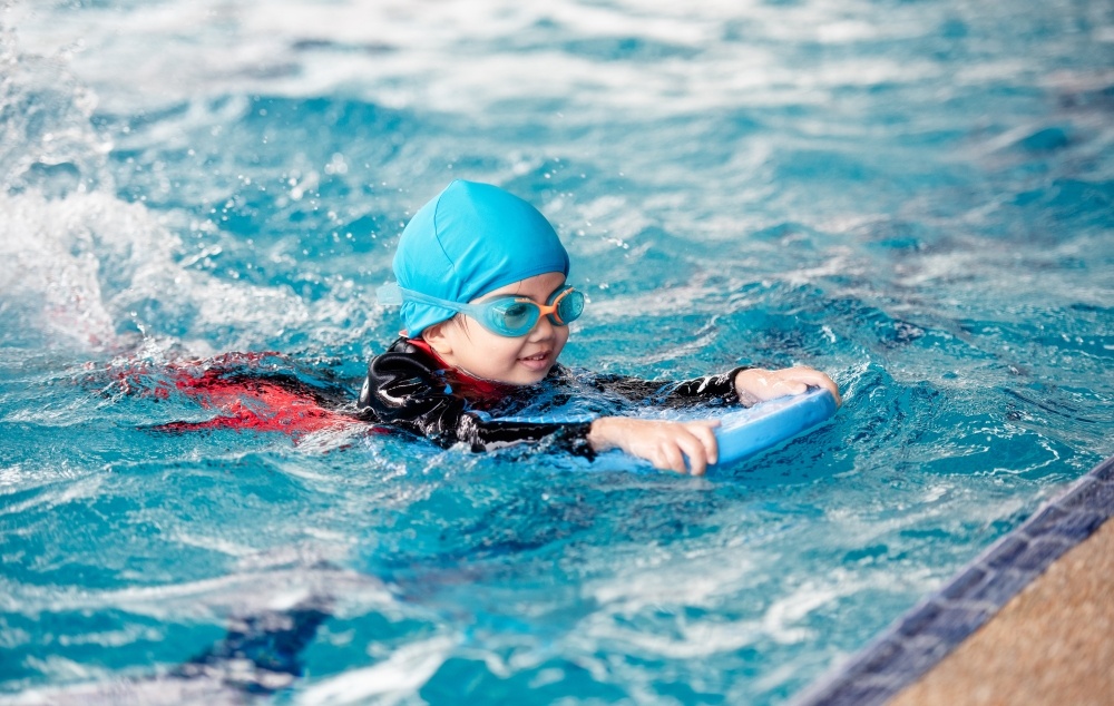 A child with a swim cap, goggles, and a kickboard swimming in a pool. 