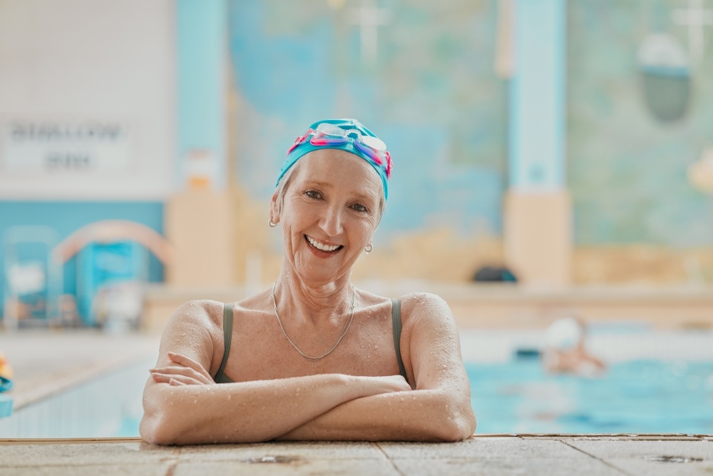 An older woman in a swim cap and goggles rests with her arms folded on the side of a pool. 