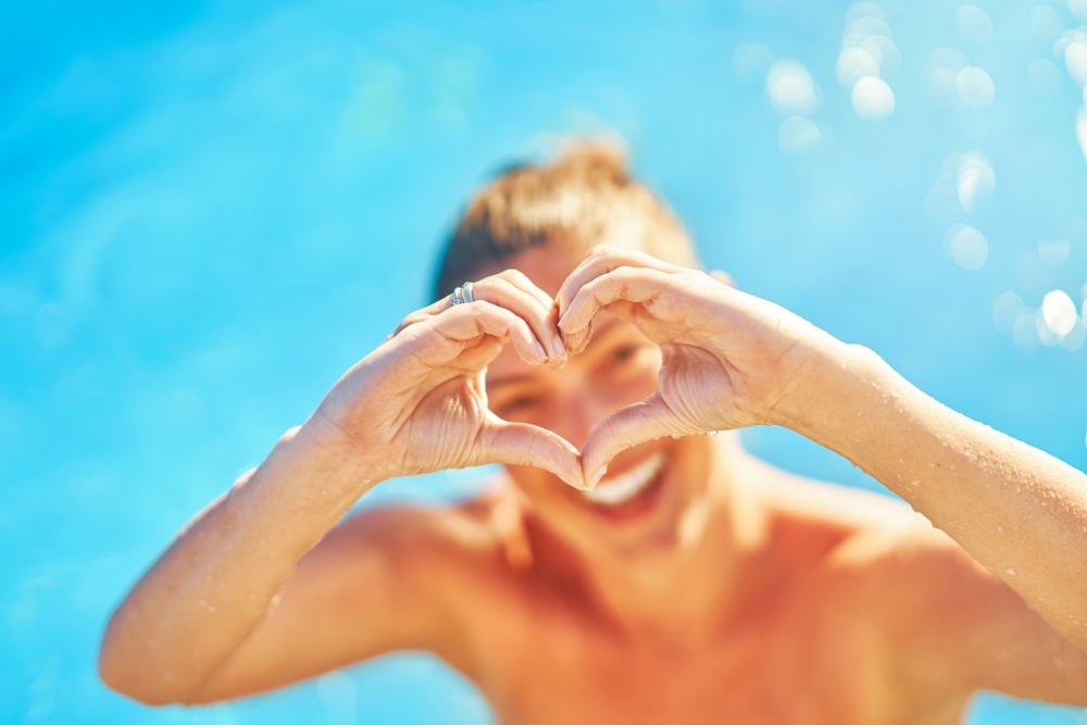 A woman makes a heart with her hands standing in front of clear pool water. Swimming is a heart healthy exercise. 