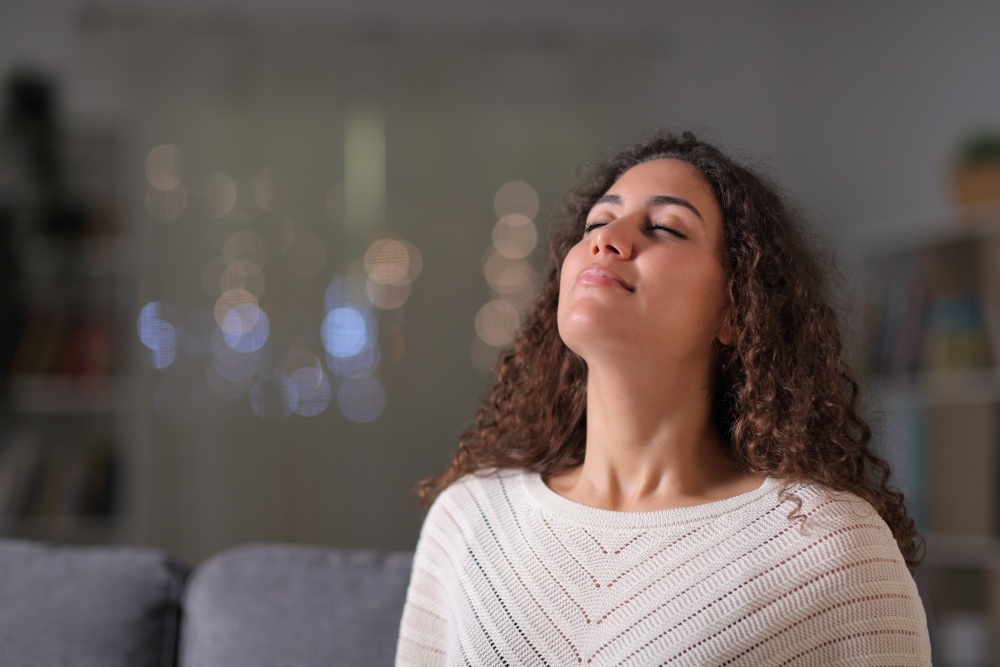 A woman sitting on a sofa looks up and smiles with her eyes closed. 