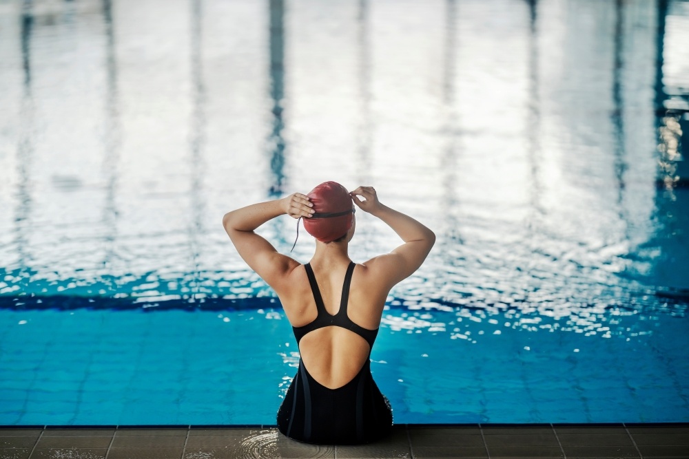 The back of a woman sitting on the edge of a pool in a bathing suit, adjusting her swim cap. 