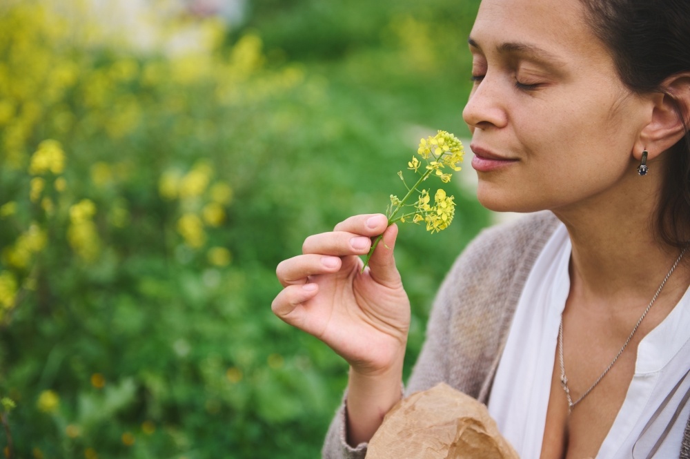 A woman takes a deep breath holding a small yellow flower to her nose in a garden. 