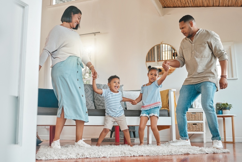 A mom, dad, and two little kids stand in a bright living room, holding hands and dancing. 