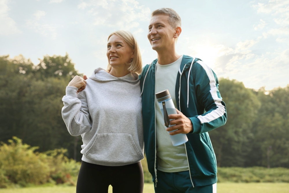 A couple in athletic clothing stand with their arms around one another, the man holding a water bottle. 
