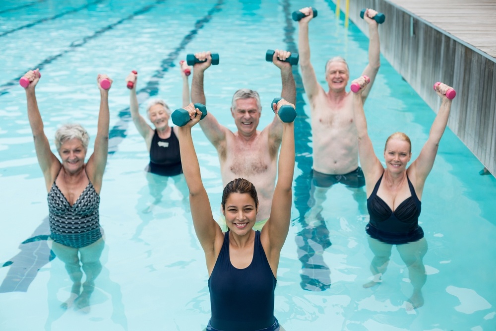 A group of adults standing in waist-high water, holding dumbbells over their head and smiling.