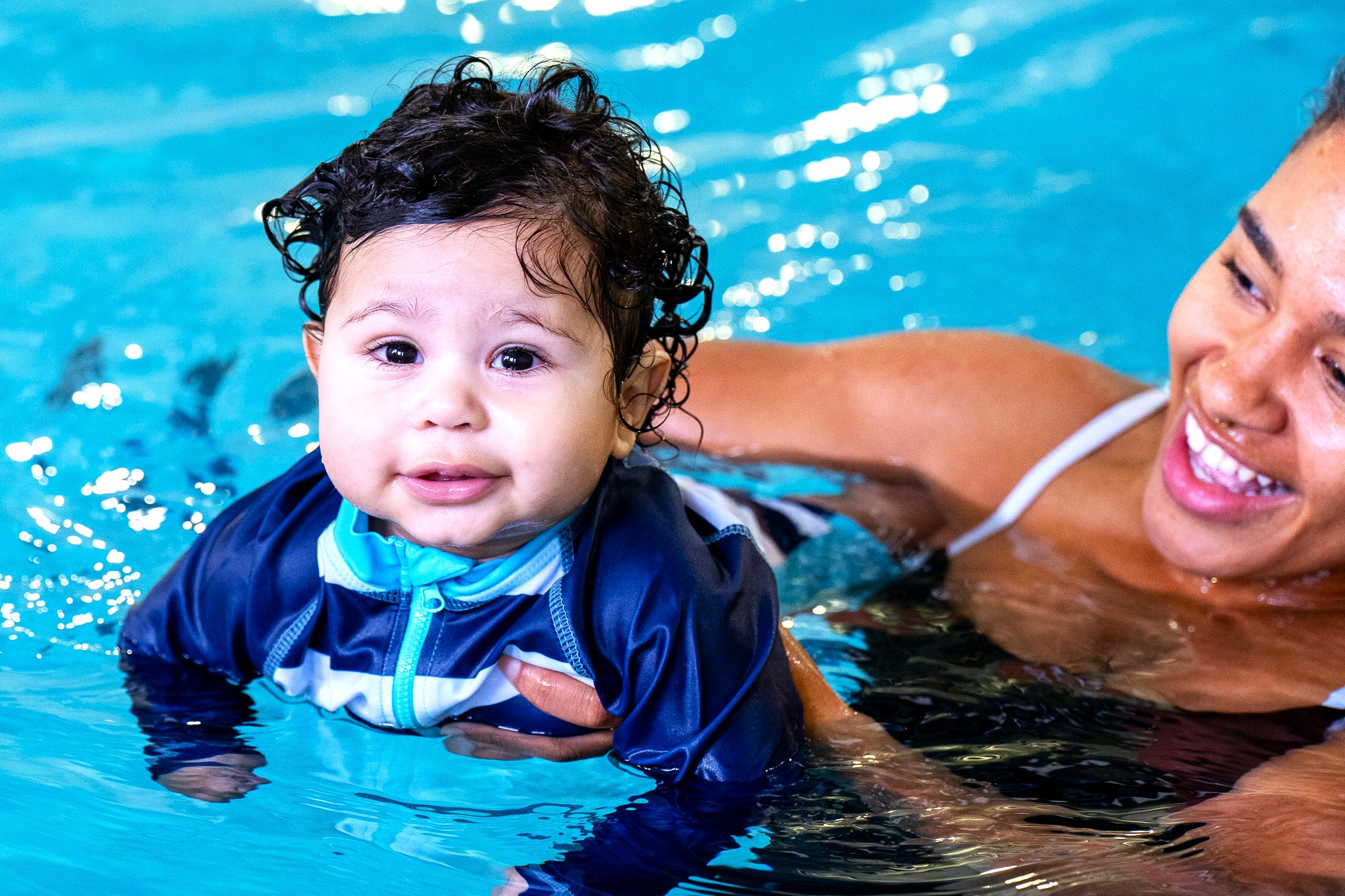 A smiling baby floats in the water supported by his mother during a swim lesson.