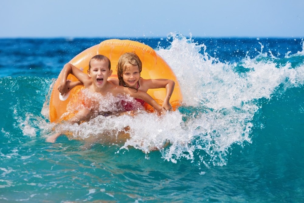 A boy and girl in a yellow inner tube ride a wave while laughing and smiling.