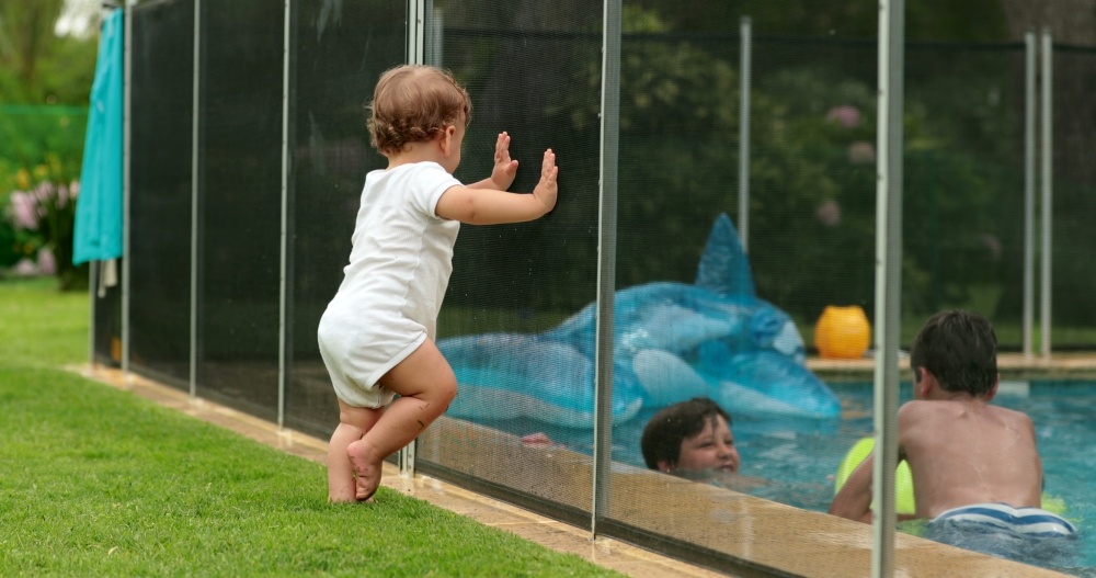 A toddler leans on a barrier fence around a pool, looking in at several kids who are swimming.