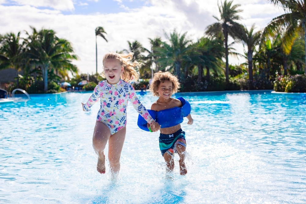 A boy and girl in bathing suits run through pool water surrounded by palm trees.