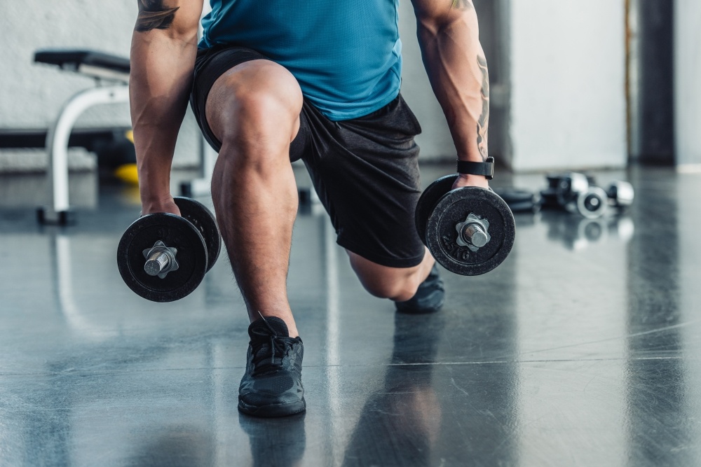 A close-up of a man's lower body as he squats, holding dumbbells at his side. 