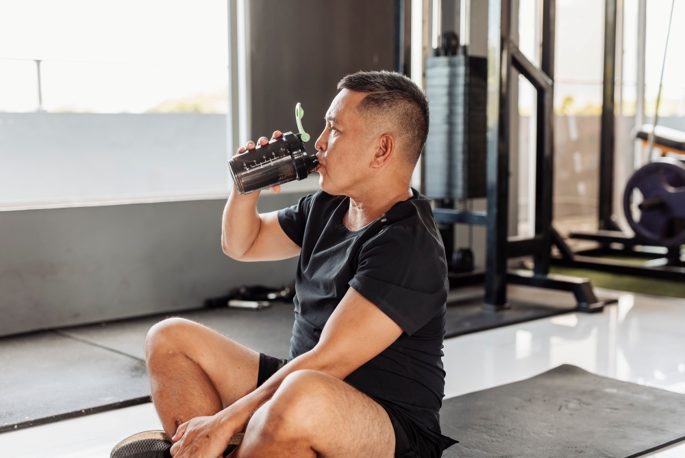 A man sits indoors on a yoga mat drinking water. 