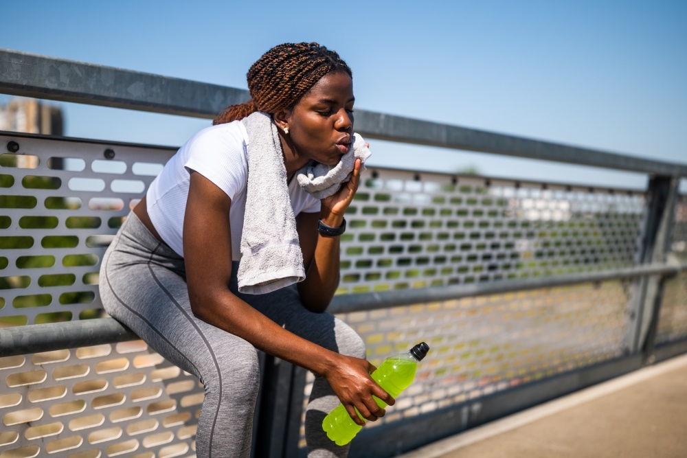 A young woman in workout gear with a bottle of electrolytes drink and a towel around her neck sits outdoors beside a metal fence, clearly overheated. 