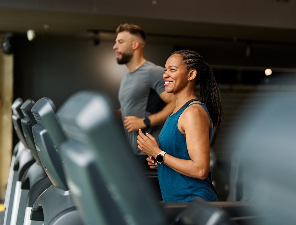 A man and woman smiling as they run on indoor treadmills beside one another. 