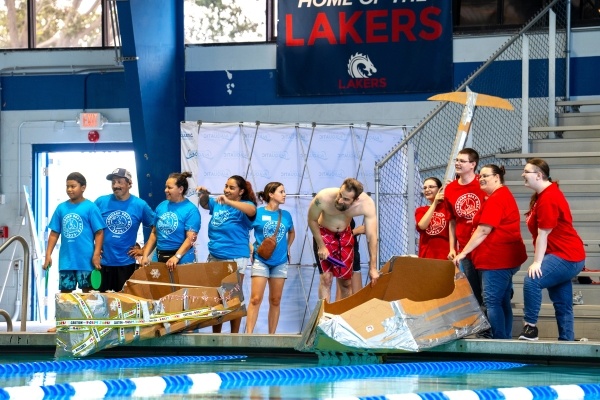 A team in blue shirts and a team in red shirts stand on the pool deck beside cardboard boats during the RAFC Cardboard Boat Race.