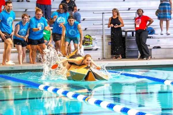 A person paddles on top of a cardboard boat in an indoor pool, beeing cheered on by her teammates at the Cardboard Boat Race at RAFC.