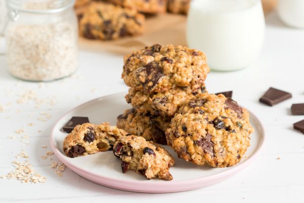 Oat cookies with dark chocolate chips served on a white plate, oats and a glass of milk in the background.