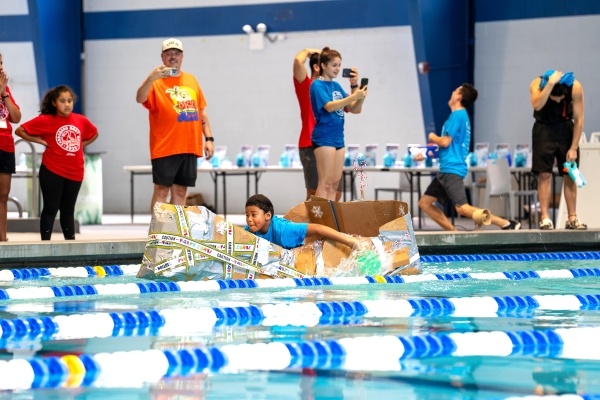 A young boy captains a cardboard boat in an indoor pool setting with other people cheering him on from the pool deck.